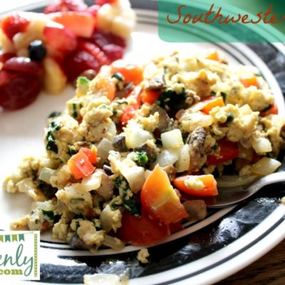 Eggs with spinach, tomatoes, and onions with fruit and black and white plate in the back round