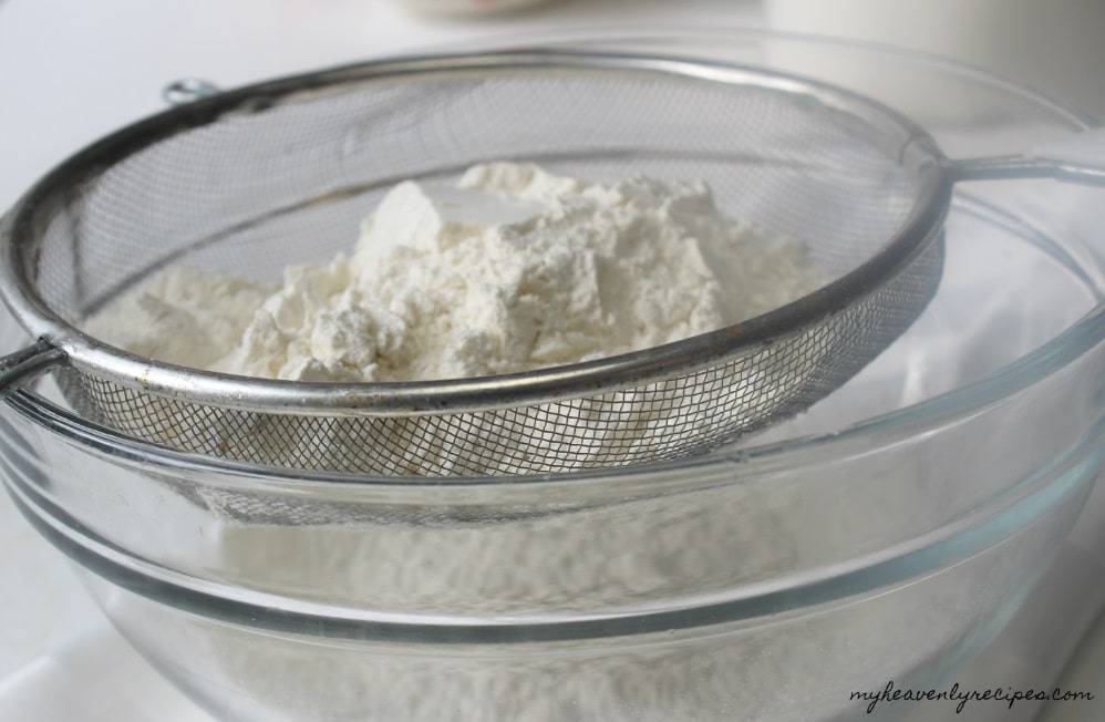 flour being sifted through a strainer into a glass mixing bowl