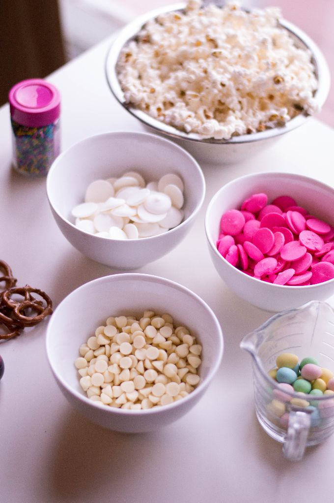 overhead shot of white table with ingredients to make sweet popcorn mix for Easter, including pastel candies, popcorn, and chocolate pieces in white and pink