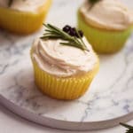 vertical image of 3 vanilla cupcakes with homemade buttercream icing on a marble platter with blueberries and rosemary sprig