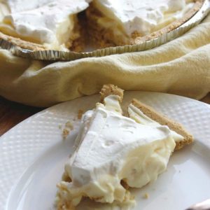 slice of no bake banana cream pie sitting on a white plate next to a fork with pie in background