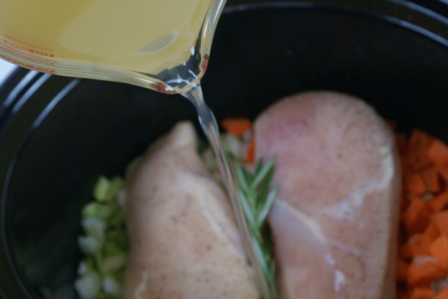 chicken broth being poured onto chicken breasts, chopped carrots, chopped celery, and rosemary